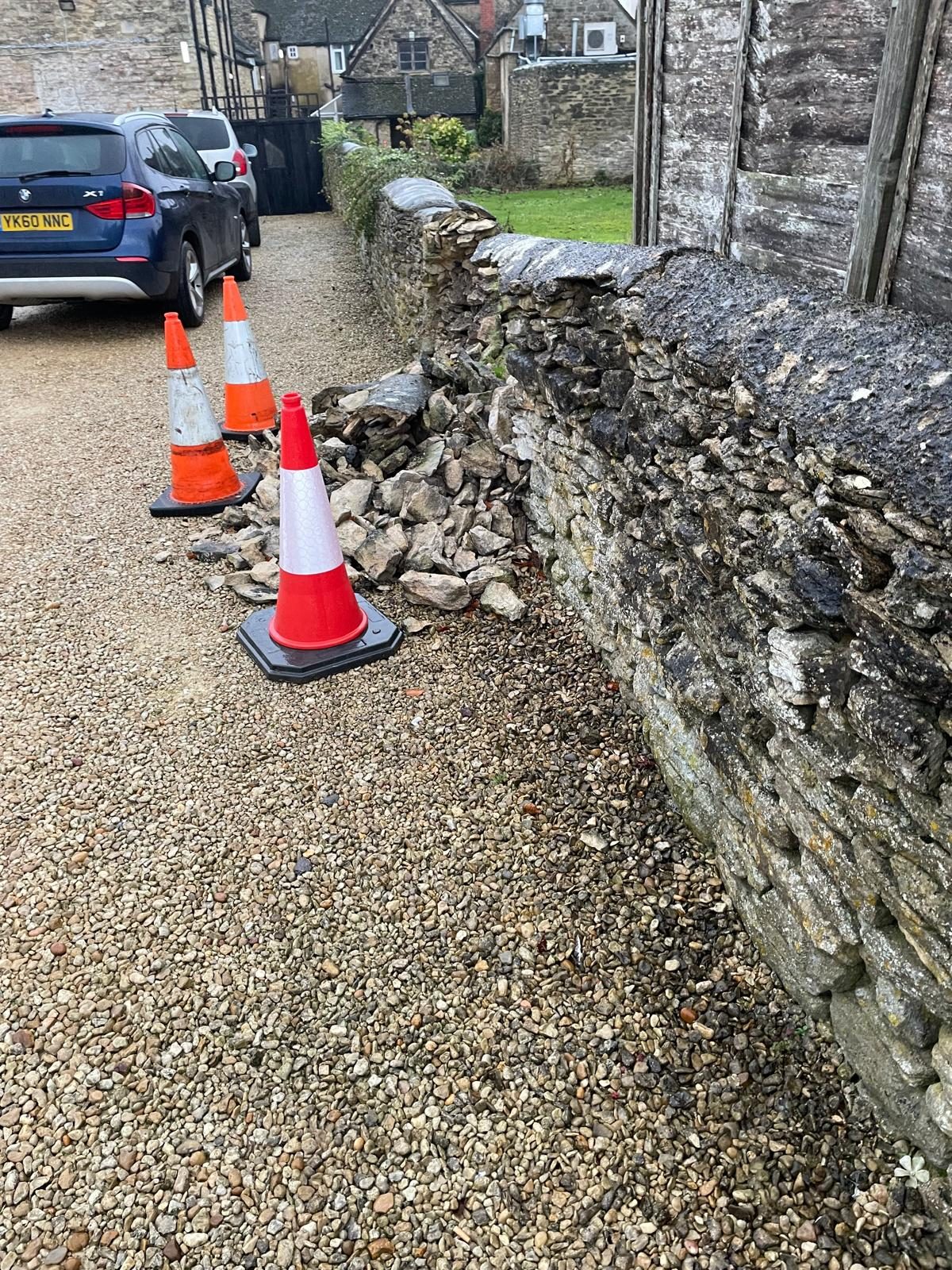 Collapsed section of dry stone wall beside a gravel drive with traffic cones